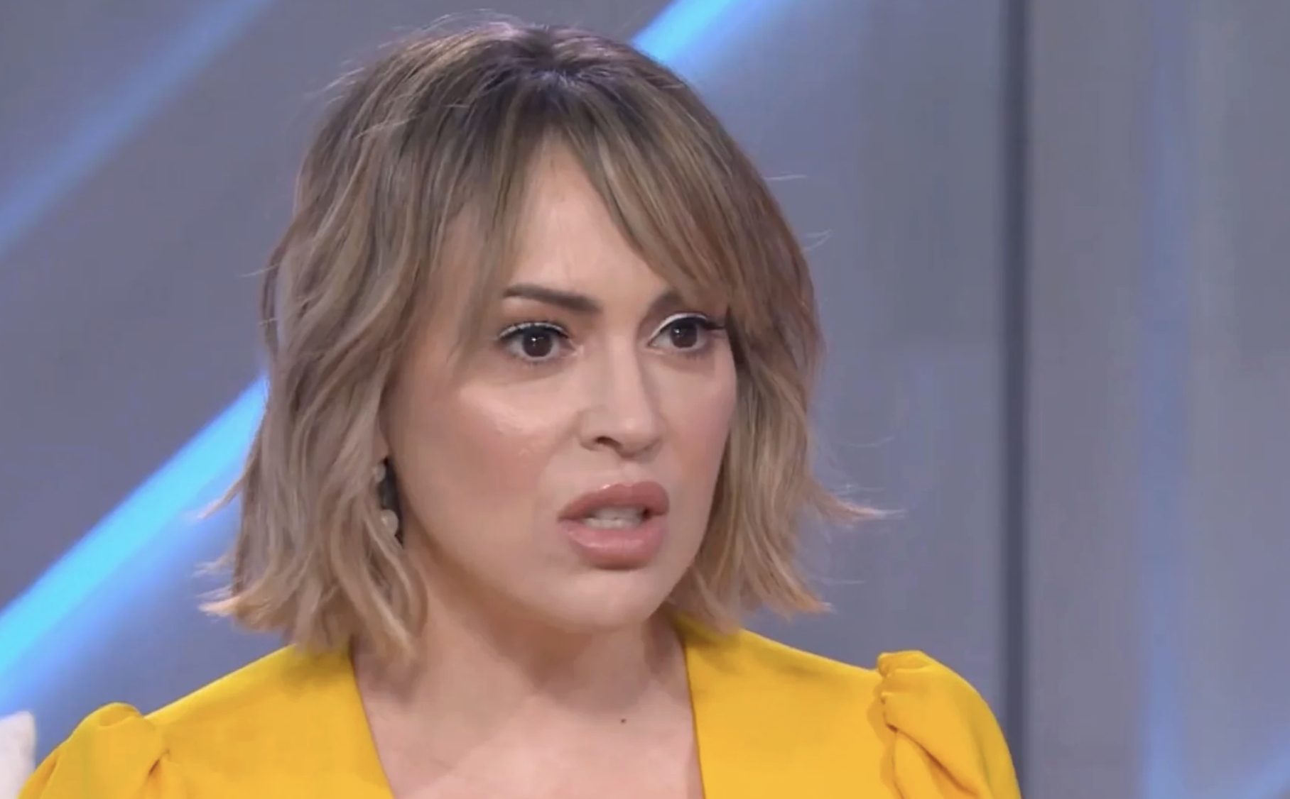 A woman with short, wavy blonde hair and a serious expression is wearing a bright yellow top. The background is out of focus with diagonal blue and gray lines.