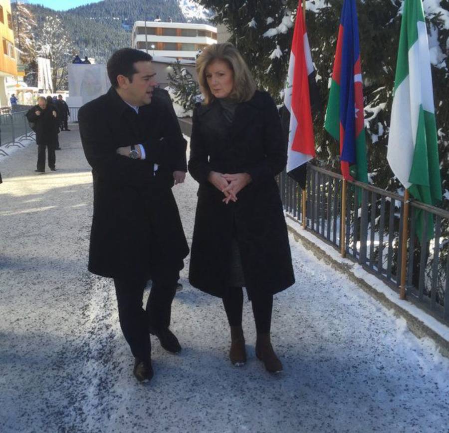Two people in winter coats walk and talk on a snowy path lined with trees and several national flags. Snow covers the ground, and mountains are visible in the background.