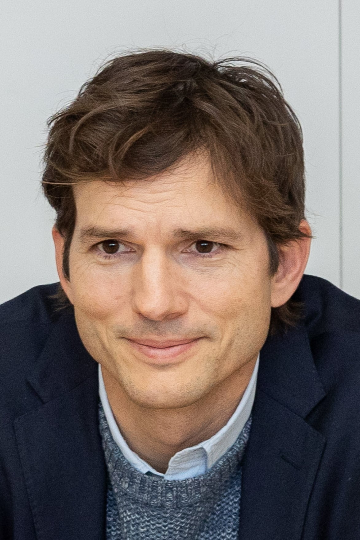 A man with short brown hair wearing a dark blazer over a gray sweater and light blue shirt is looking slightly to the side and smiling softly, with a neutral background behind him.