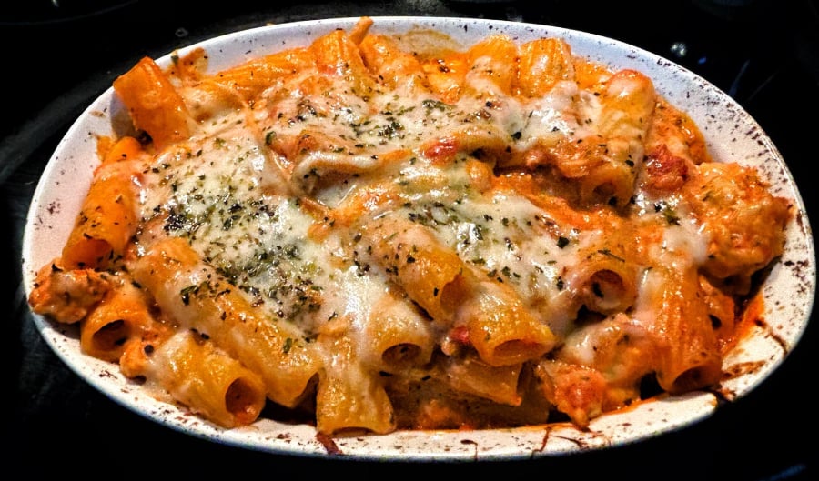 Baked pasta dish in a white oval casserole topped with melted cheese, herbs, and visible pieces of rigatoni and sauce.