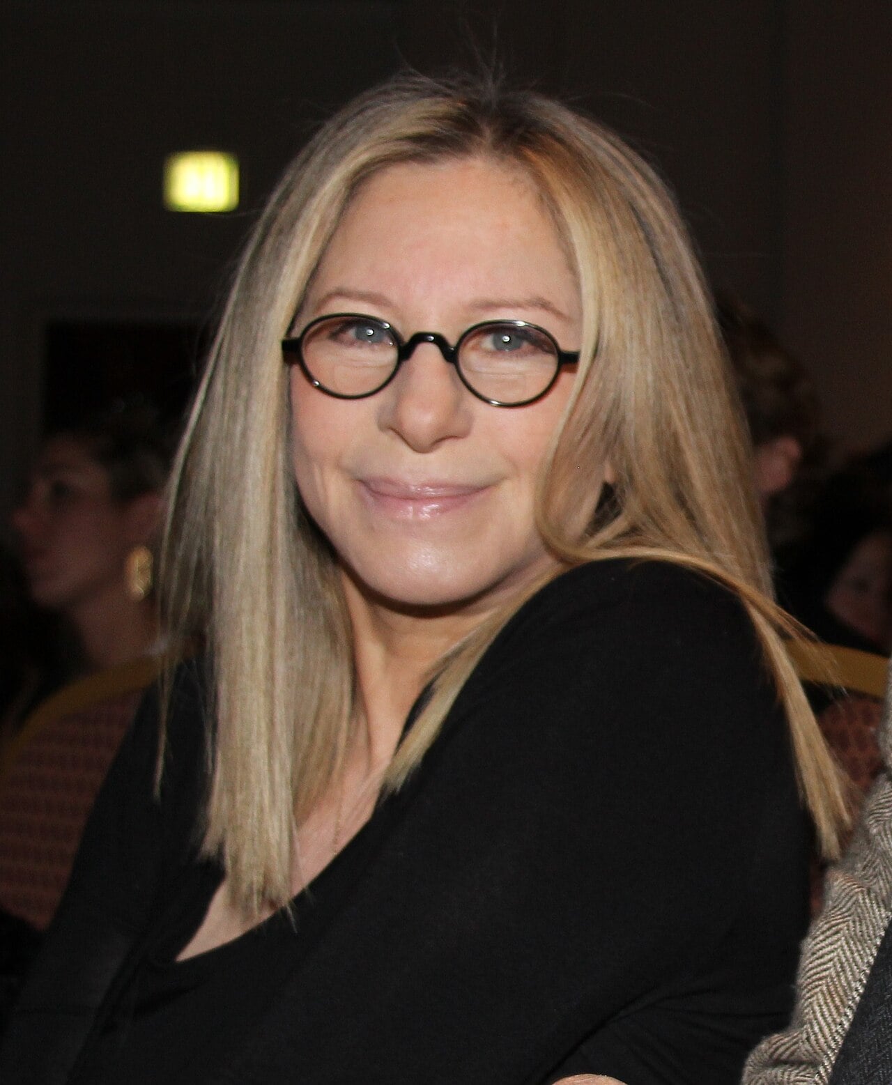A person with straight, shoulder-length blonde hair and round black glasses smiles while seated indoors, wearing a black top. The background is softly blurred with other people visible.