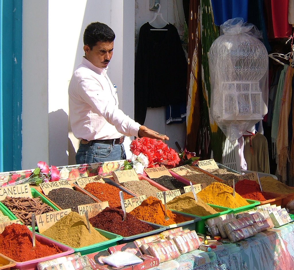 A vendor gestures toward colorful piles of spices displayed on a market stall. Each spice is labeled, and various goods hang in the background, including clothes and a birdcage covered in plastic.