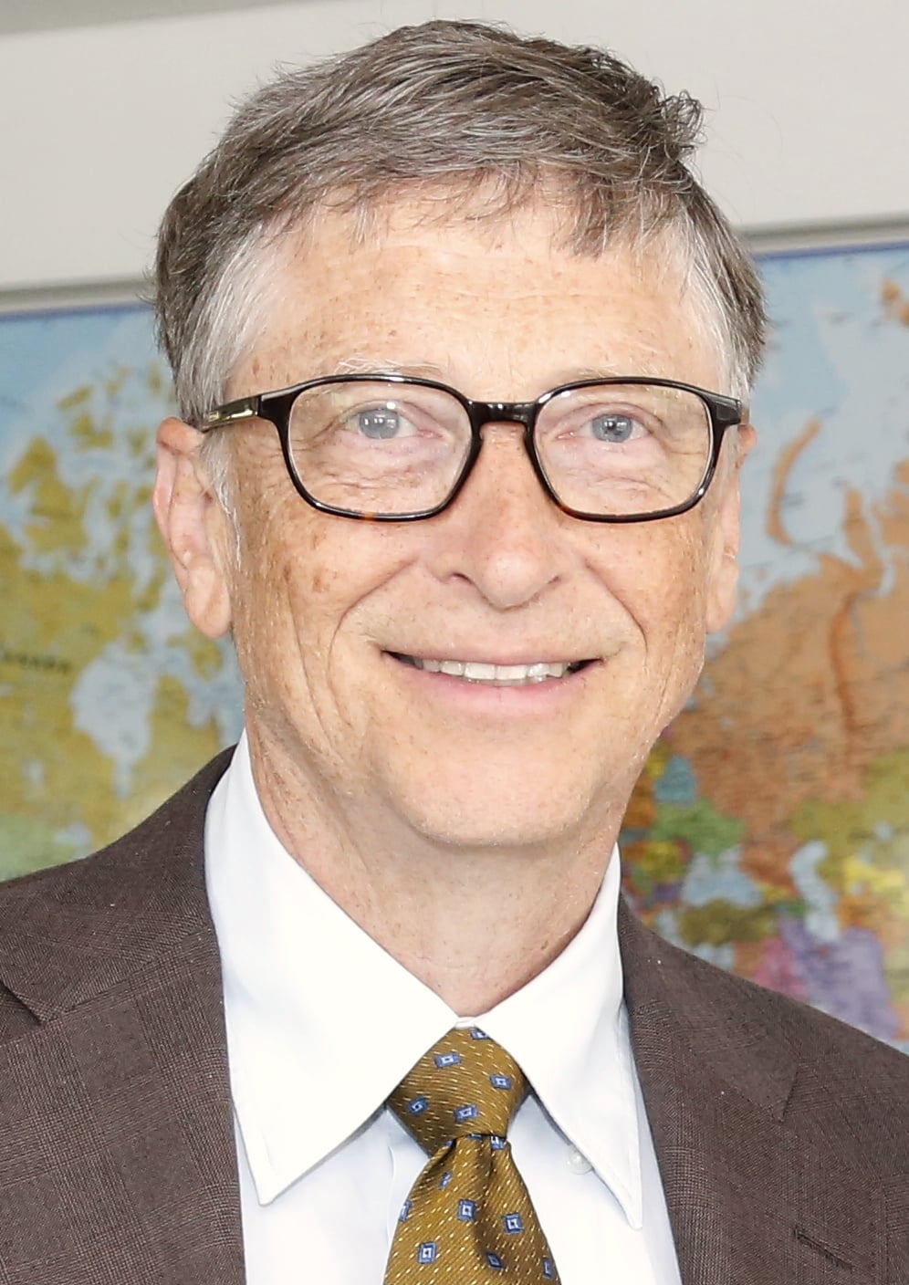 A man with gray hair and glasses, wearing a brown suit, white shirt, and patterned tie, smiling in front of a world map.