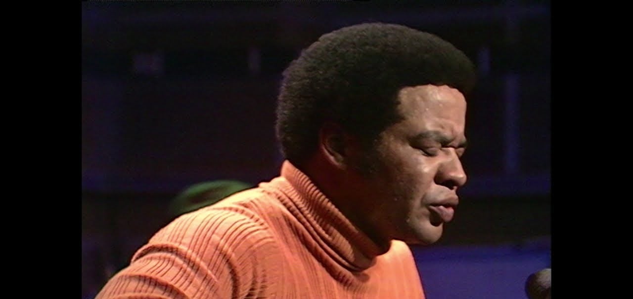A man with a short afro hairstyle wearing an orange turtleneck sweater is singing passionately with his eyes closed against a dark, blurred background.