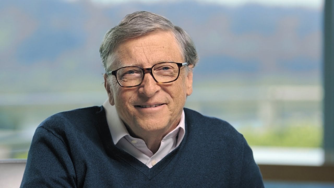 An older man with gray hair and glasses smiles at the camera. He is wearing a navy sweater over a collared shirt, seated indoors with a blurred outdoor background visible through a window.