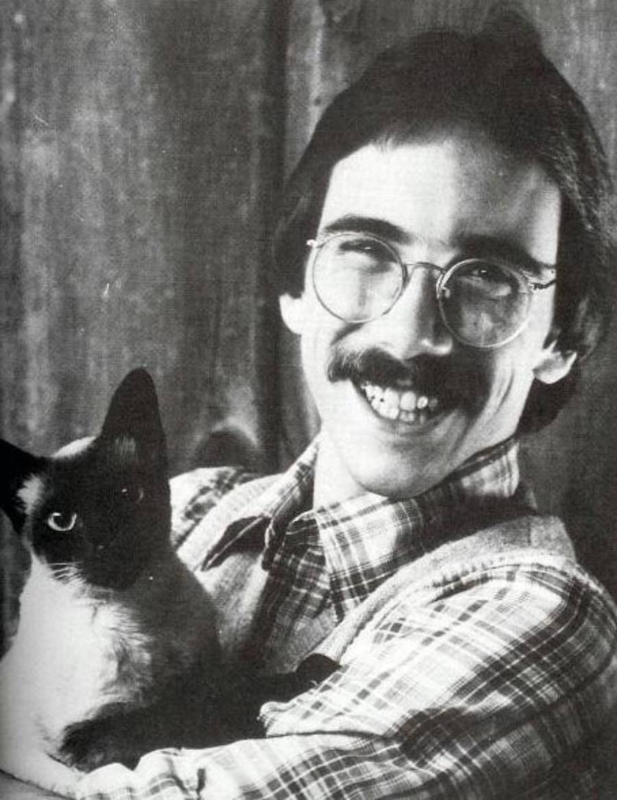 A smiling man with glasses and a mustache, wearing a plaid shirt, holds a Siamese cat in his arms. The photo is black and white.