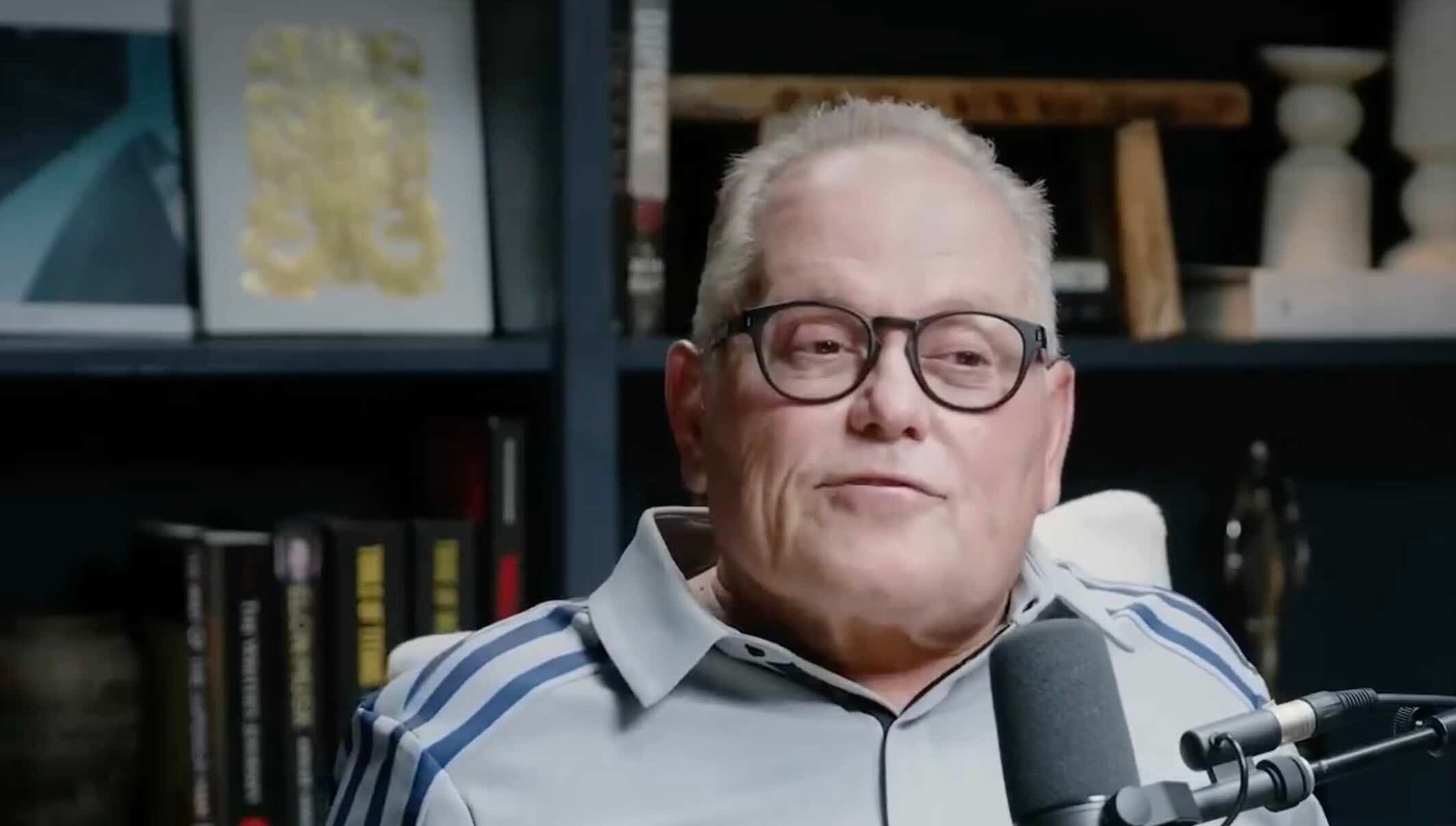 An older man with short gray hair and glasses sits in front of a microphone, wearing a light gray shirt with blue stripes, with books and decorative items on shelves in the background.