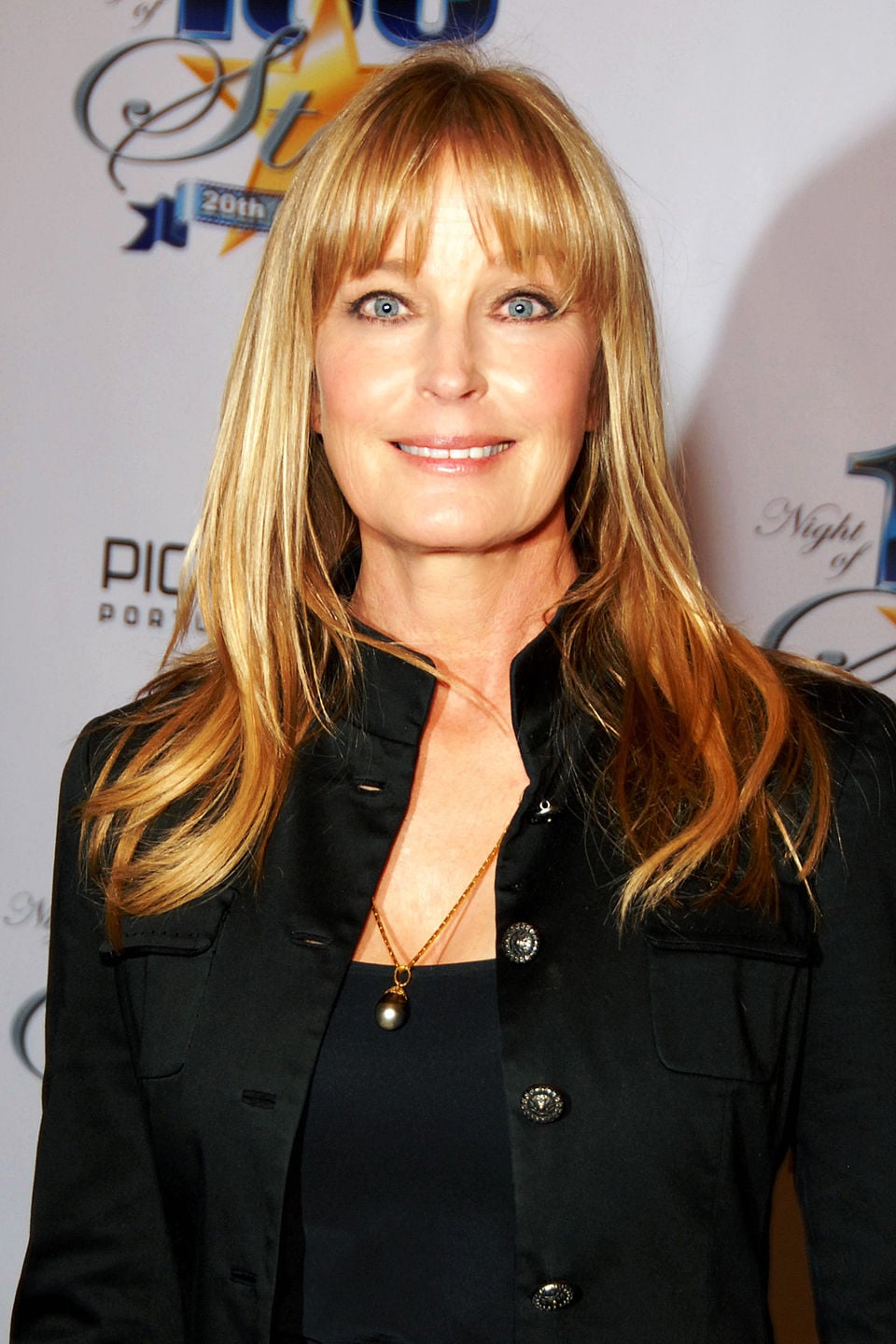 A woman with long blonde hair and bangs, wearing a black jacket and a pearl necklace, smiles while standing in front of a white backdrop with event logos and text.