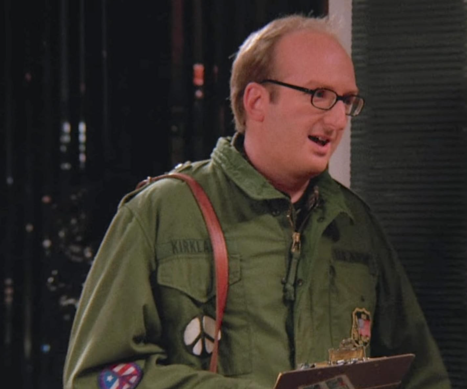 A man with glasses and thinning hair wears a green jacket with patches and a shoulder strap, holding a clipboard and standing indoors, speaking or smiling.