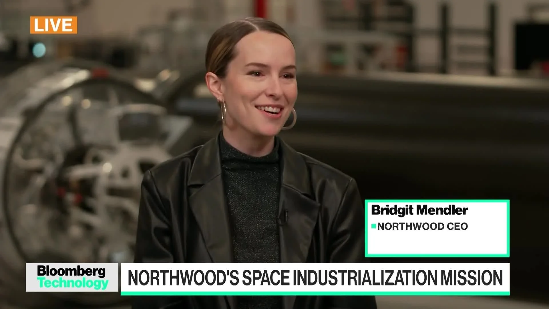 A woman, identified as Bridgit Mendler, Northwood CEO, is smiling during an interview on Bloomberg Technology. The screen headline reads: "Northwood's Space Industrialization Mission." Industrial equipment is blurred in the background.