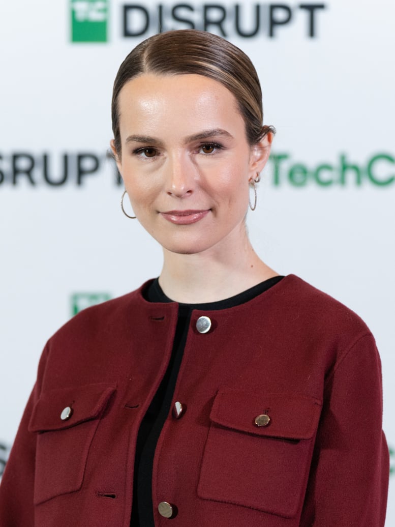 A person with slicked-back brown hair, wearing hoop earrings and a dark red jacket, stands in front of a backdrop with the words "TechCrunch Disrupt.