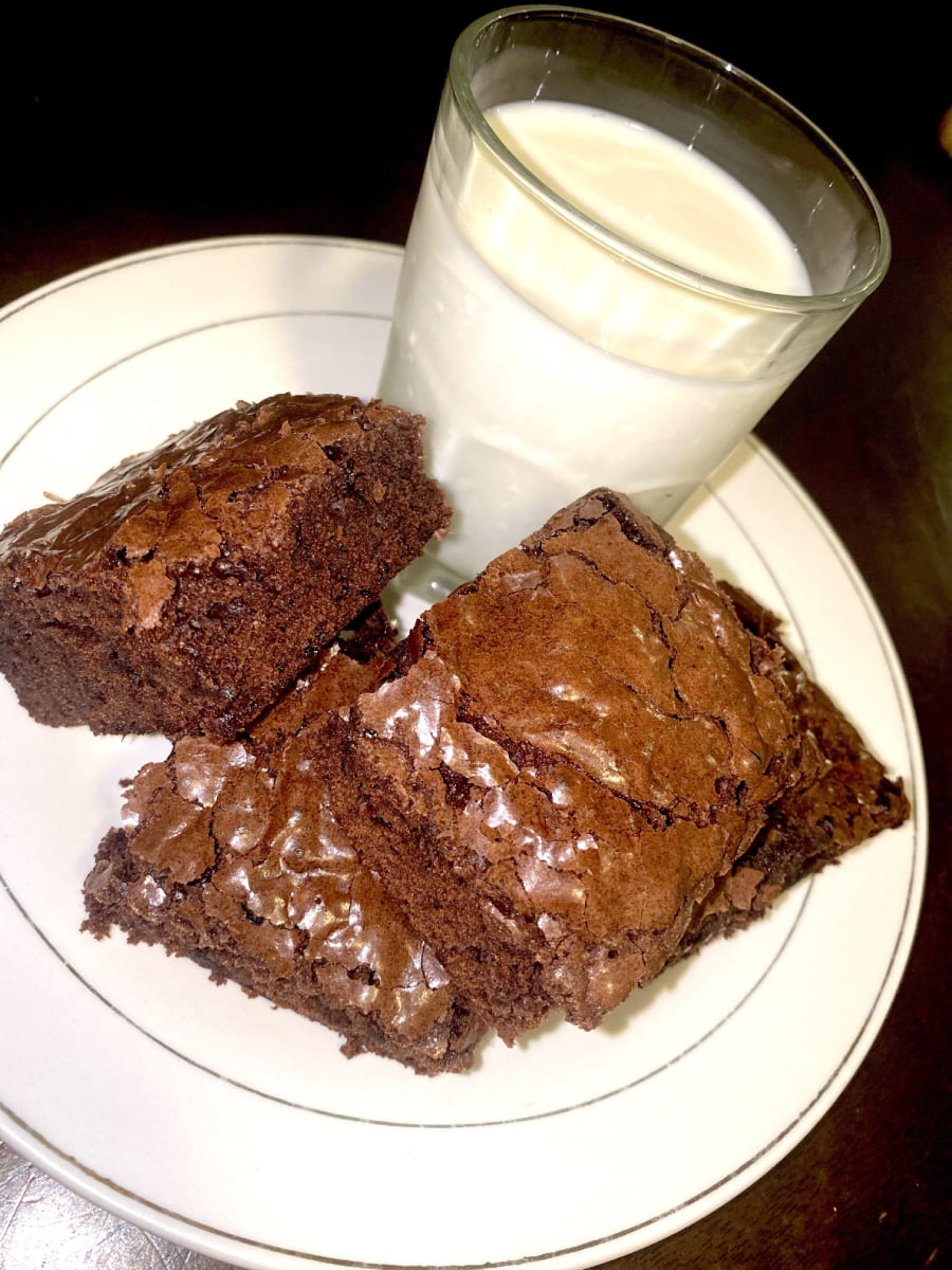 A plate with three chocolate brownies and a glass of milk placed on it, set against a dark background.