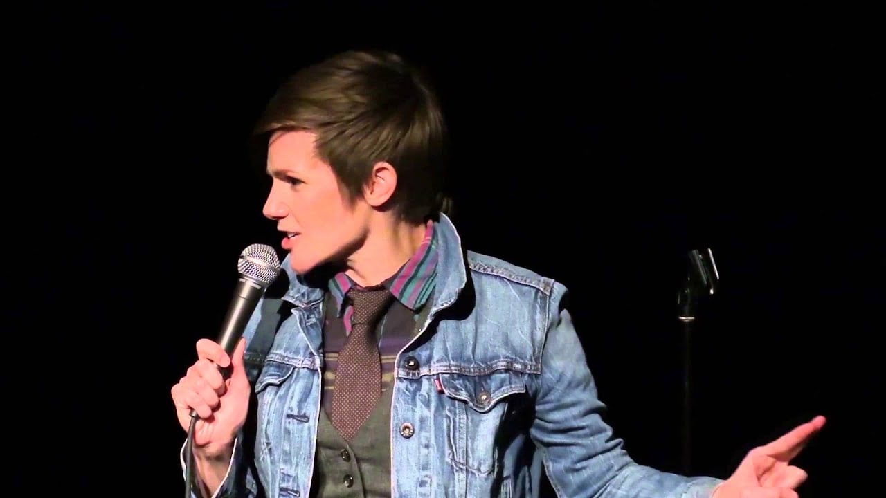 A person with short brown hair, wearing a denim jacket, shirt, and tie, holds a microphone and gestures while performing on stage against a black background.