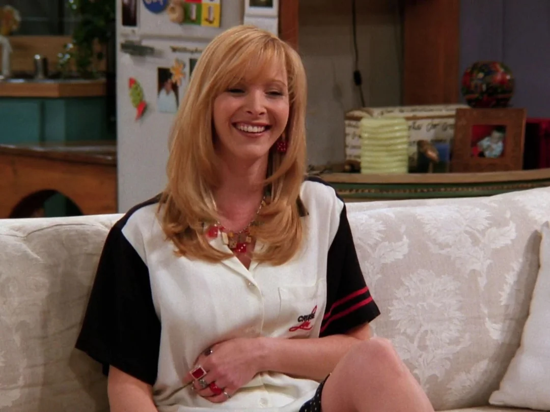 A woman with blonde hair sits smiling on a light-colored couch. She is wearing a white shirt with black sleeves, red jewelry, and has her hand resting on her lap. A kitchen and refrigerator are visible in the background.