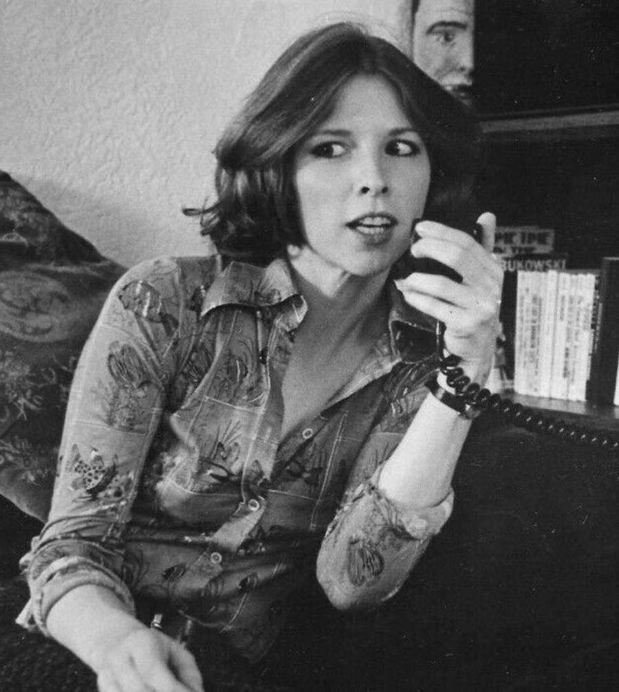 A woman with shoulder-length hair sits indoors, holding a corded microphone or intercom. She wears a patterned button-up shirt and looks slightly to the side, appearing to speak or listen intently. Books are visible in the background.