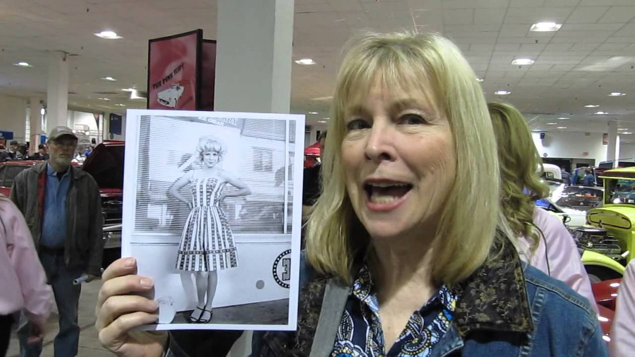 A smiling woman with blonde hair holds up a black-and-white photo of another woman in a patterned dress at an indoor car show, with classic cars and people in the background.