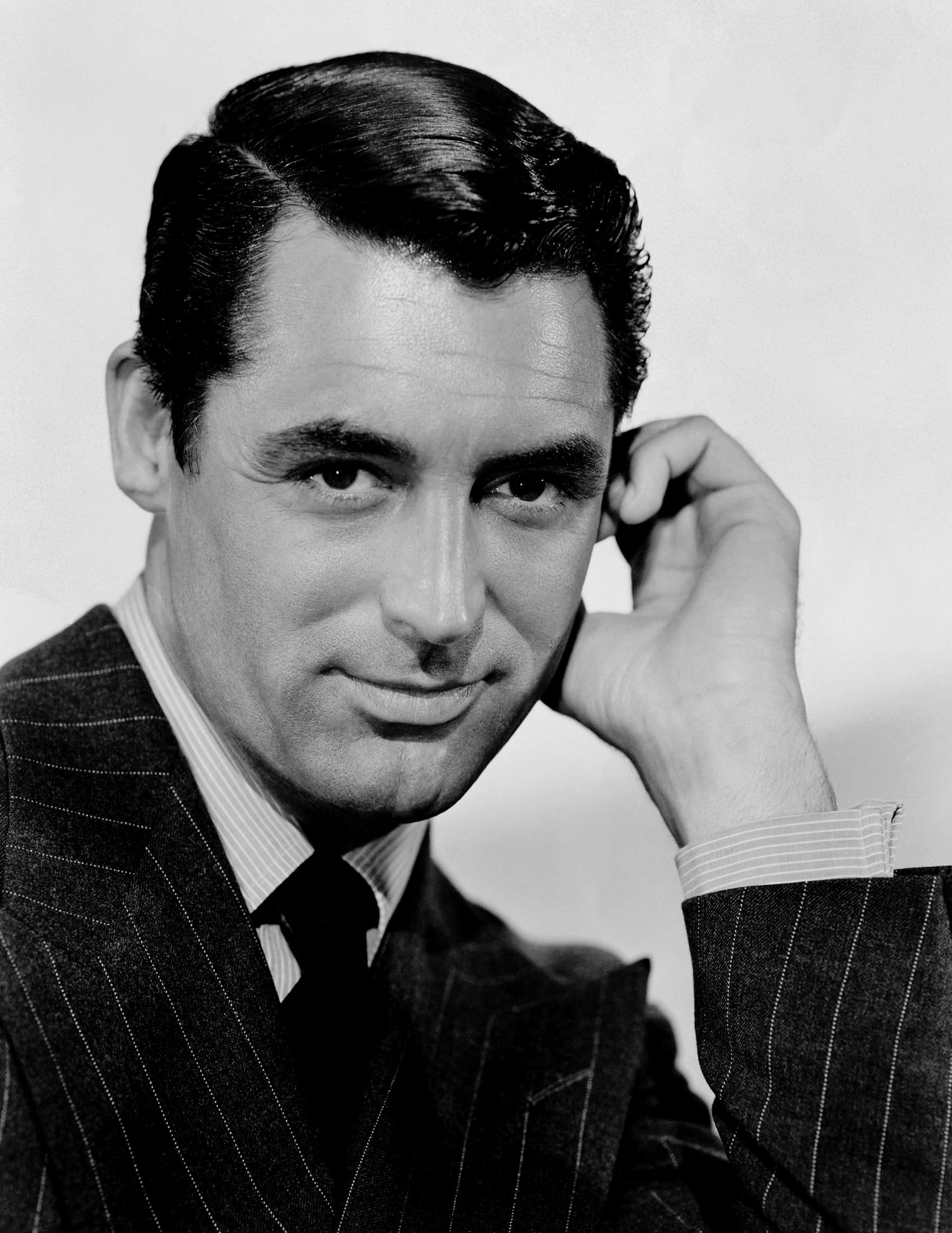 A man in a pinstripe suit with neatly combed dark hair rests his head on one hand and looks confidently at the camera against a plain background. The image is in black and white.
