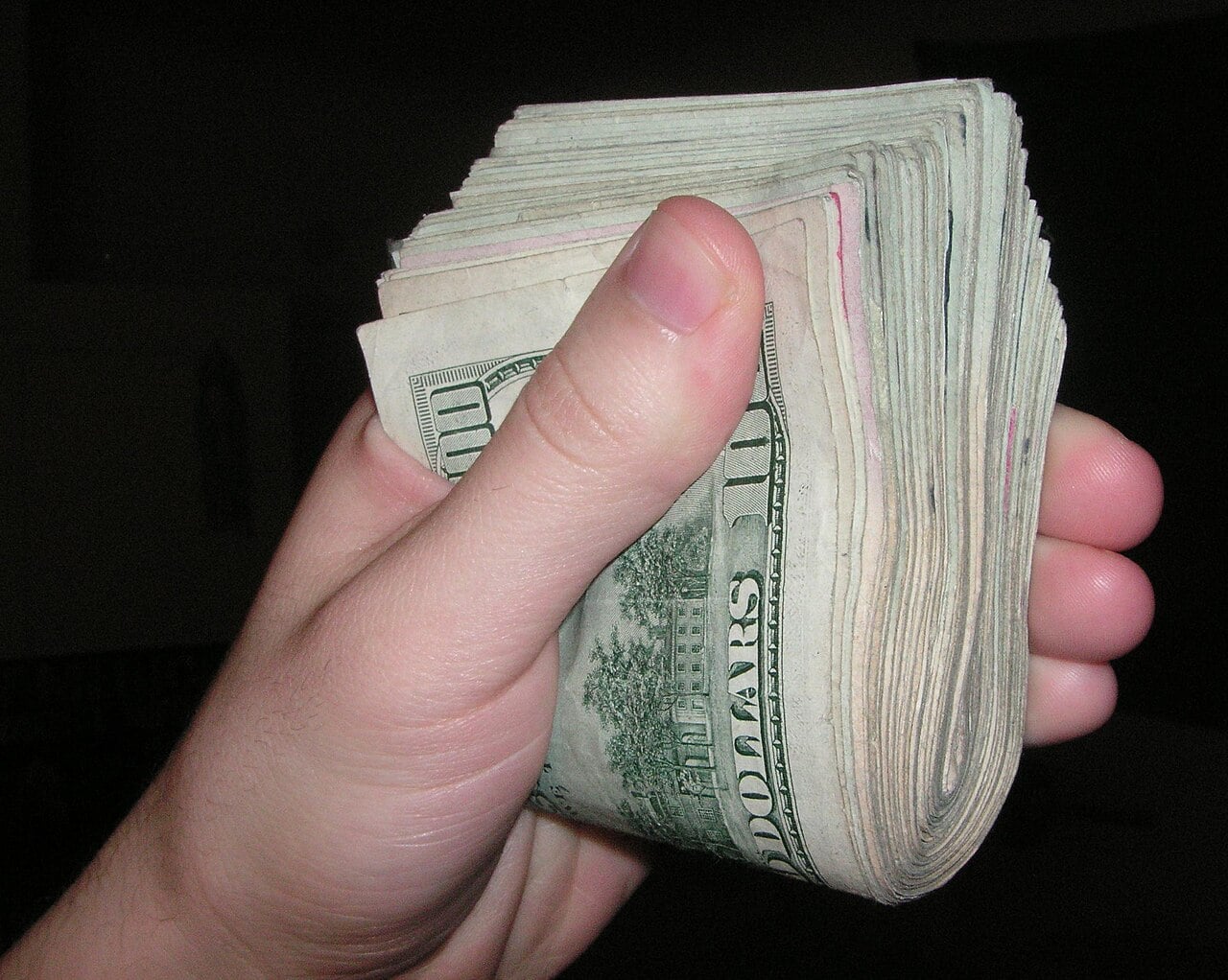 A hand holding a thick stack of U.S. hundred-dollar bills, fanned out and gripped firmly against a dark background.