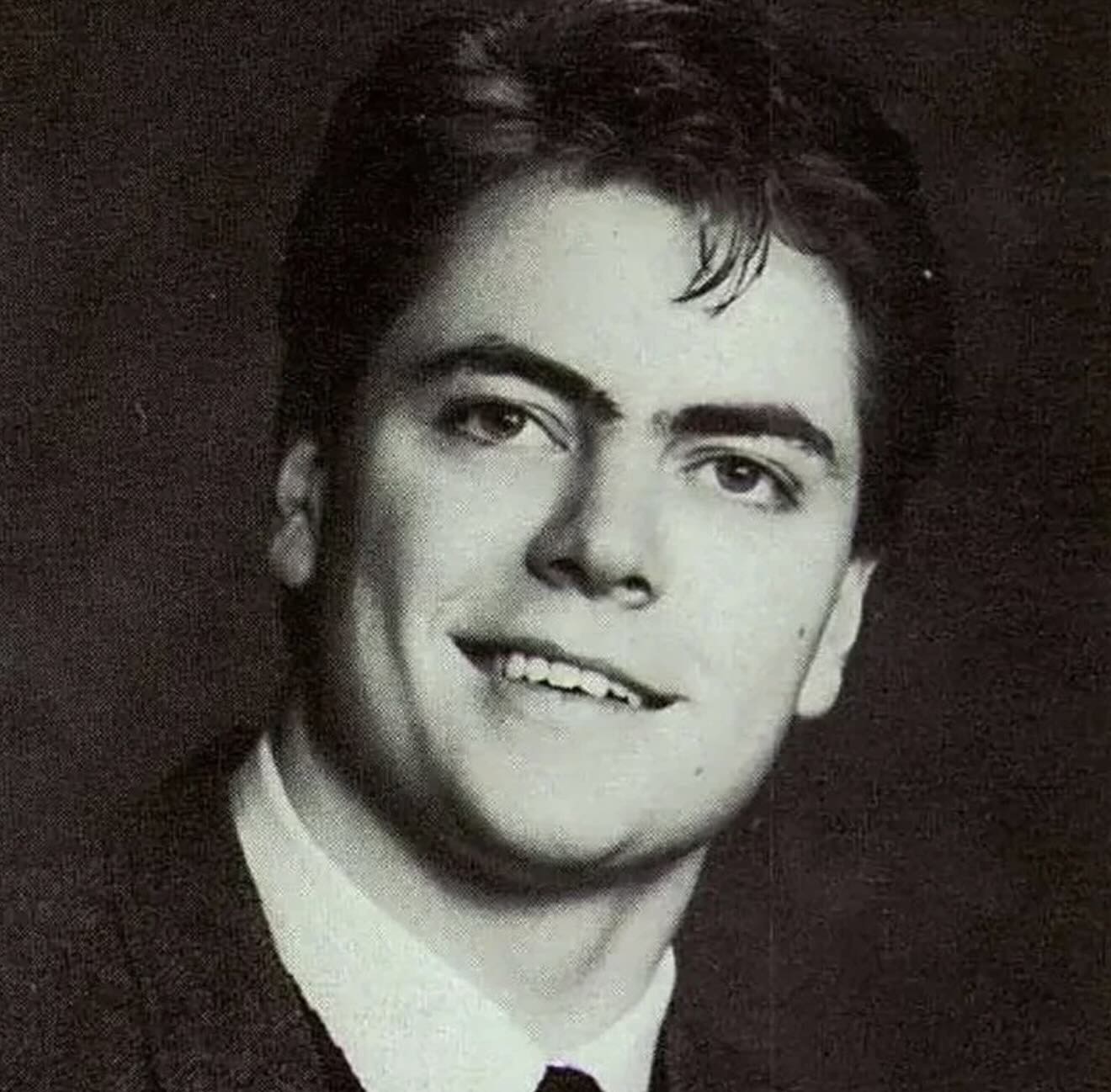 A young man in a black-and-white portrait, wearing a suit and tie. He has dark hair, combed back, and is smiling slightly while looking just off camera. The background is plain and dark.