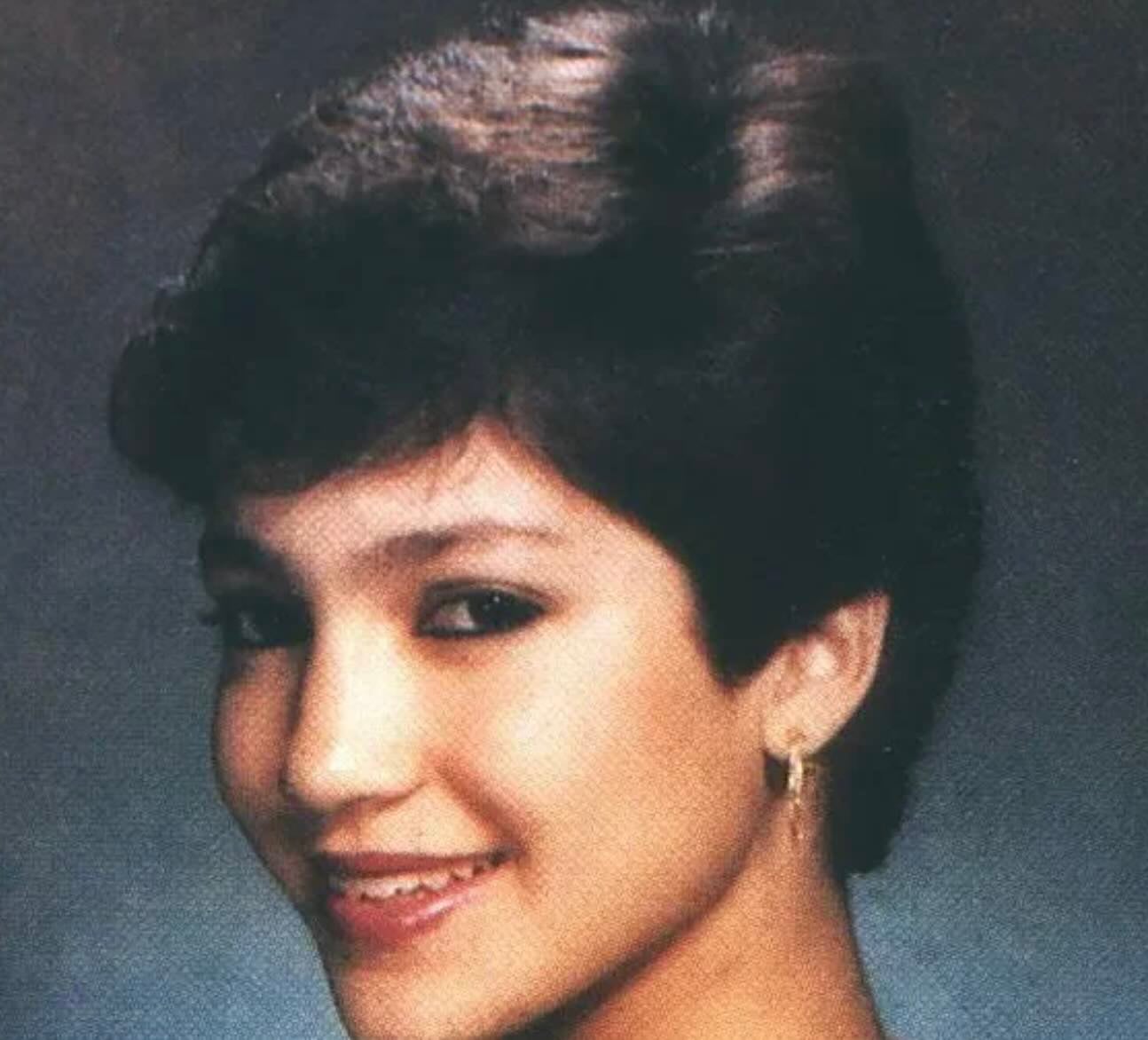 A young woman with short, dark hair smiles at the camera. She is wearing small hoop earrings and a subtle makeup look, posing against a dark, studio-style background.