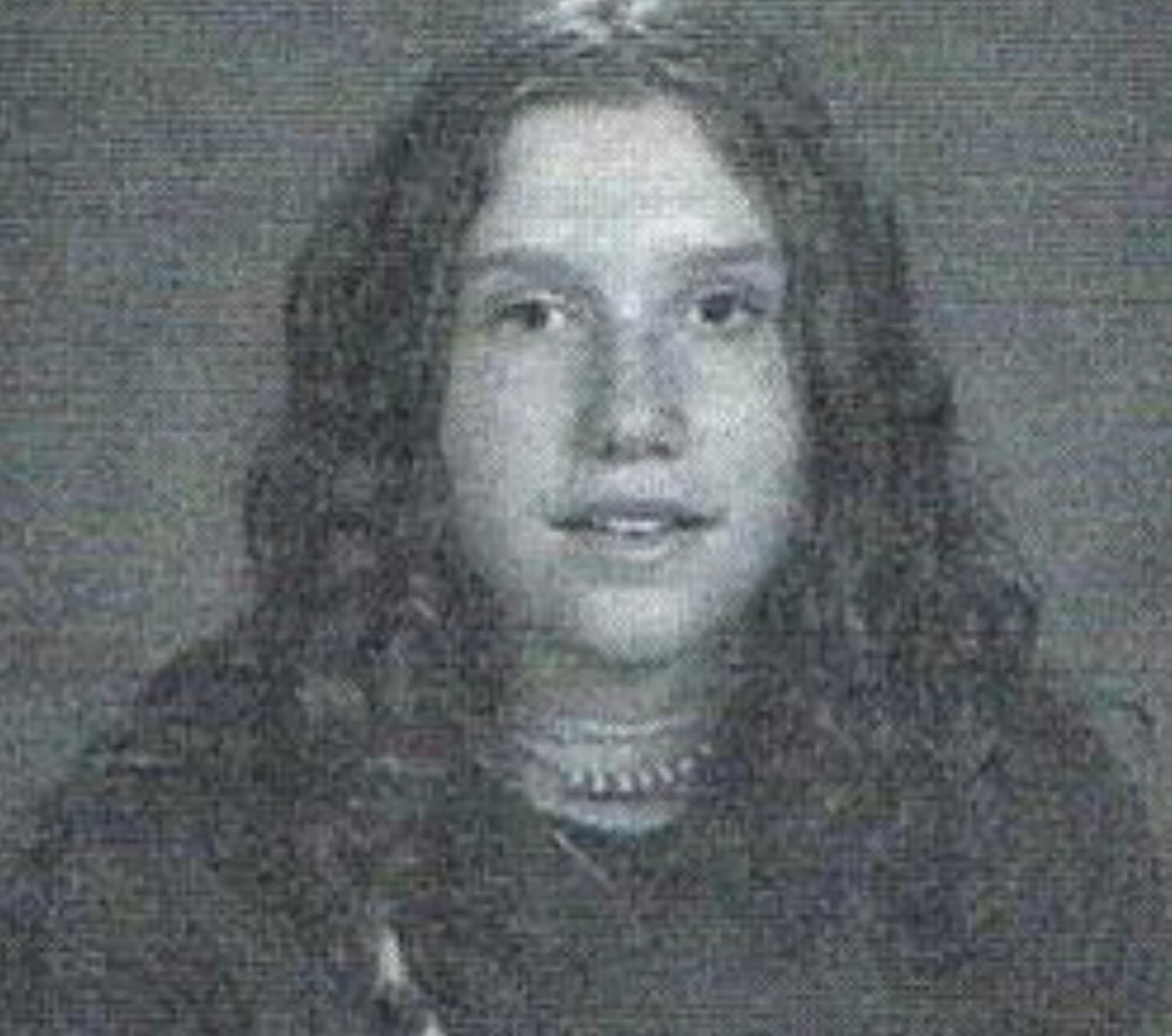 A black and white portrait of a young person with long, wavy hair, wearing a layered necklace and a dark top, looking directly at the camera with a neutral expression.