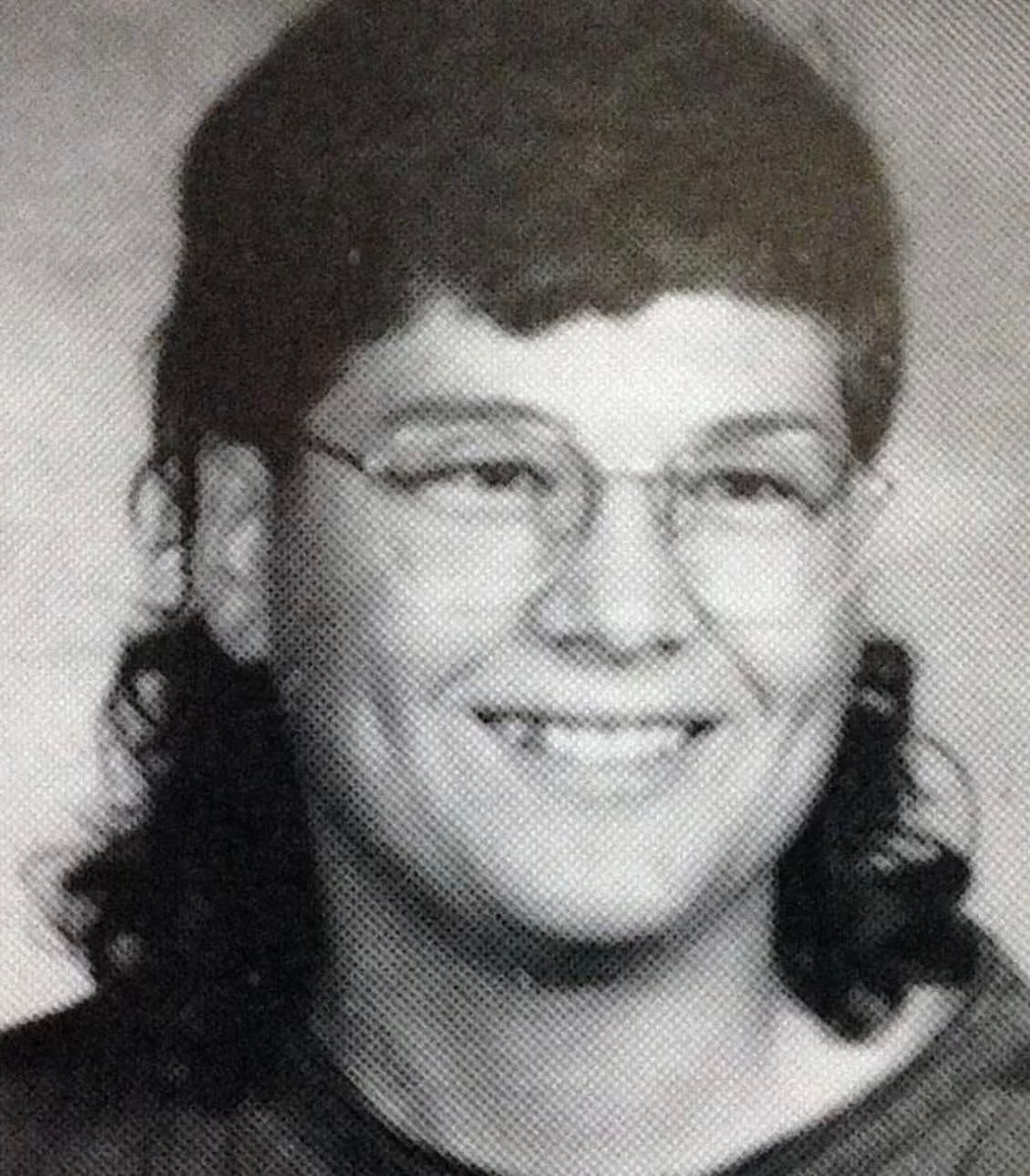 Black-and-white yearbook photo of a person with short, curly hair on top and longer hair in the back, wearing glasses and smiling at the camera.