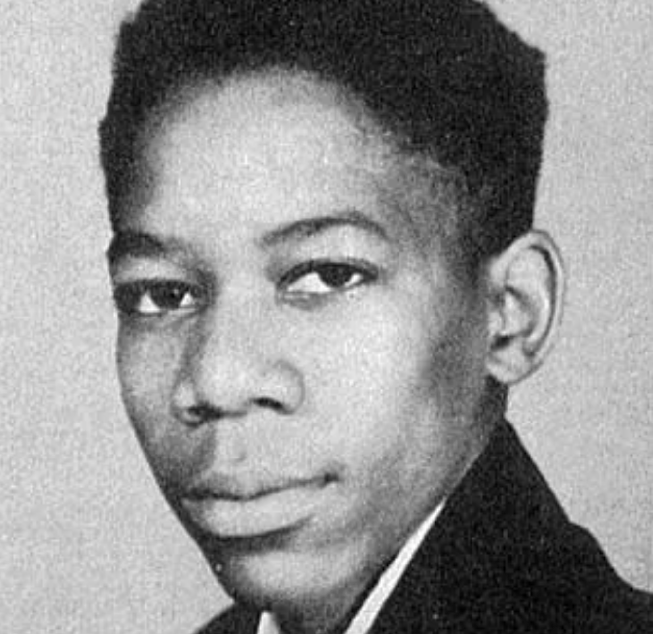 Black and white portrait of a young person with short, textured hair, wearing a suit jacket and looking slightly to the side with a neutral expression.
