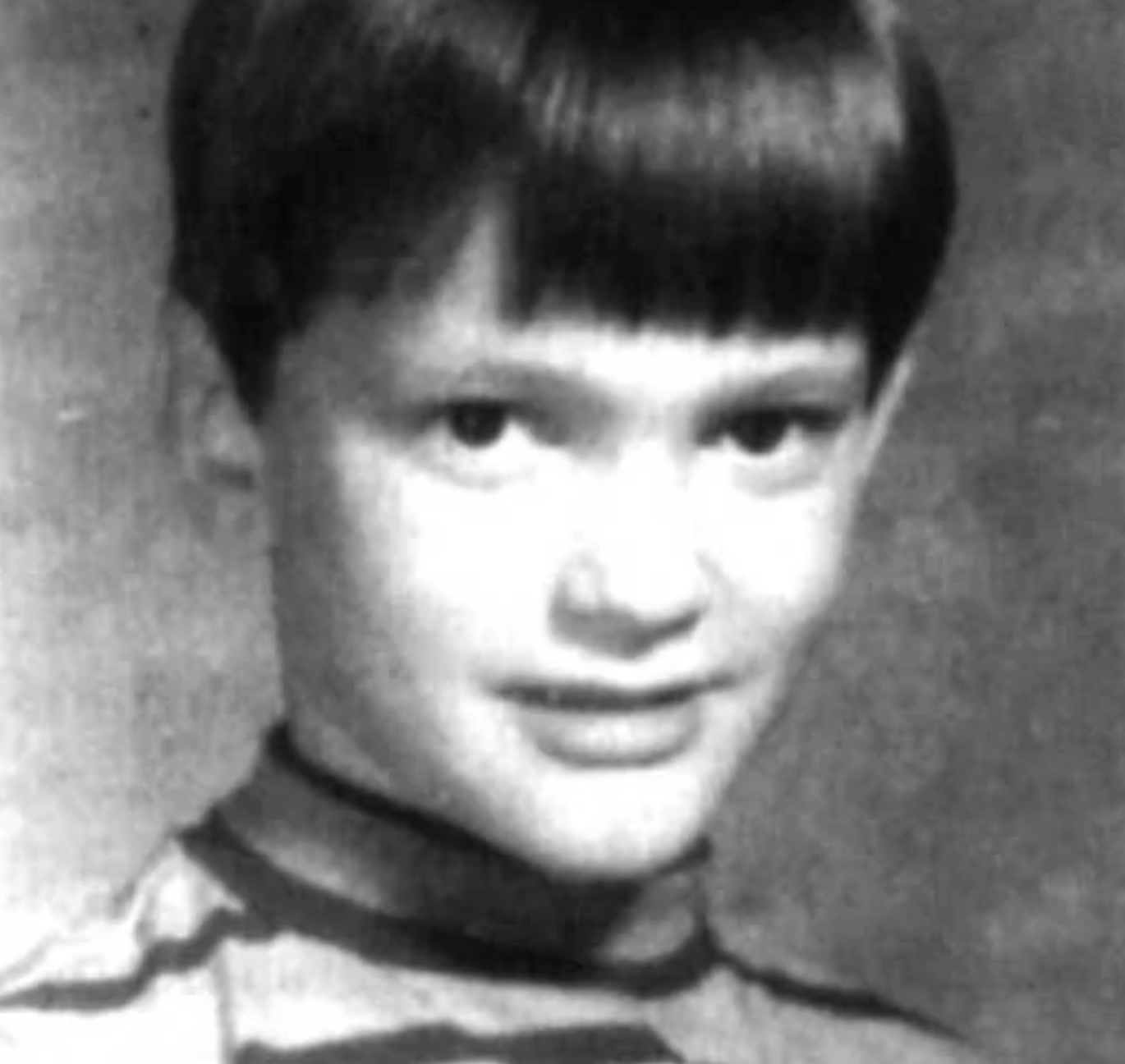 Black and white portrait of a young child with short, straight dark hair and bangs, wearing a striped shirt and looking slightly sideways at the camera with a subtle smile.