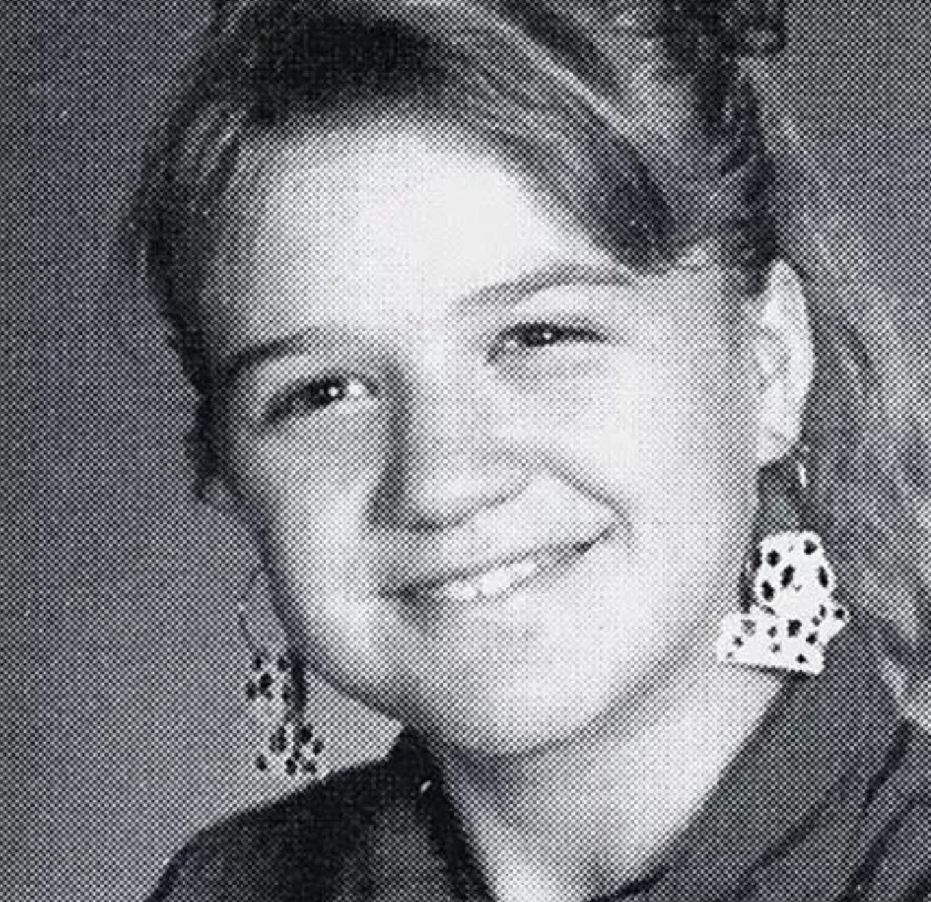 A young girl with light hair smiles at the camera in a black-and-white portrait. She is wearing large, patterned earrings and a collared shirt. The photo background is plain and neutral.