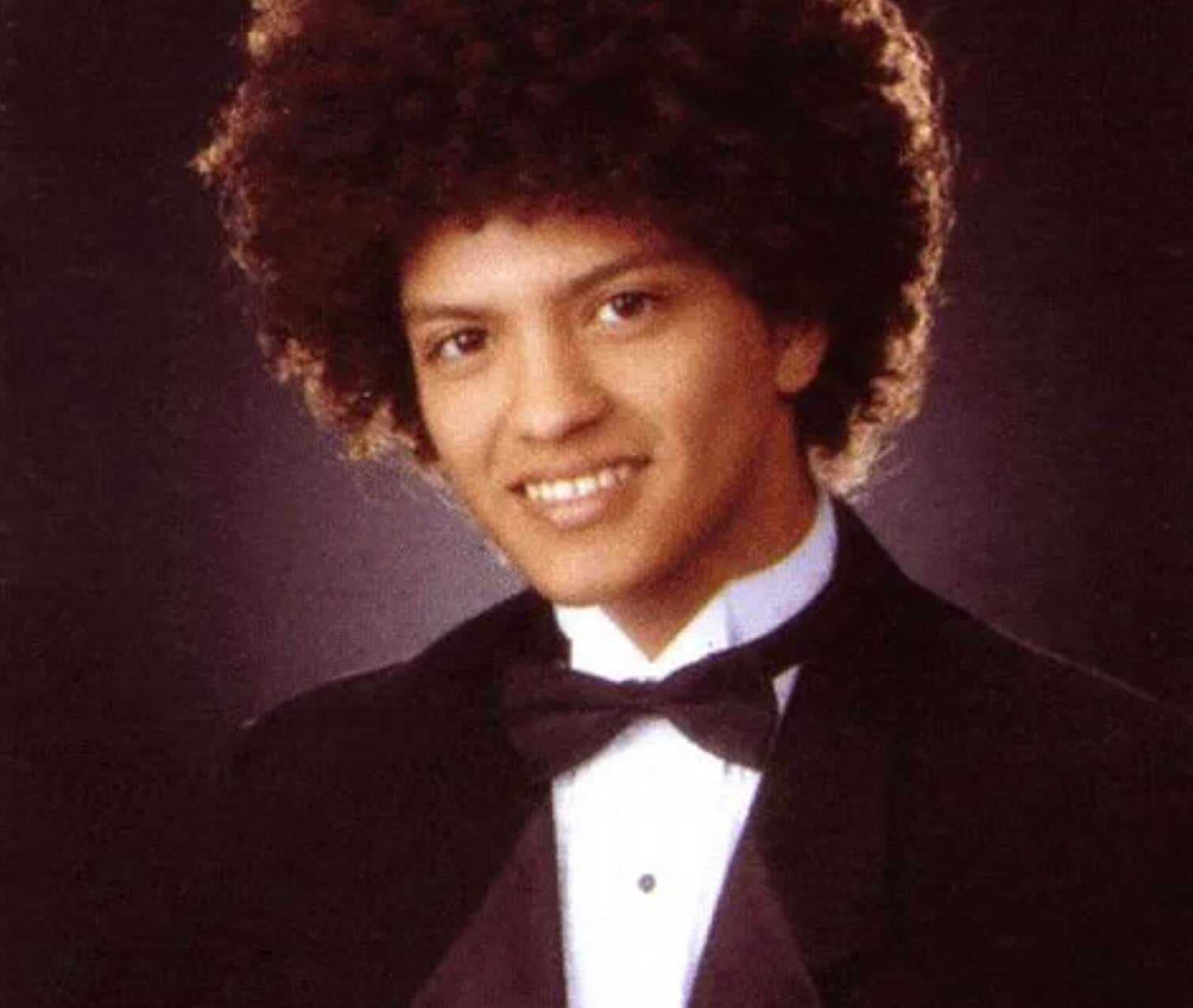 A young man with curly hair is wearing a black tuxedo and bow tie, smiling at the camera against a dark, softly lit background in a formal portrait style.