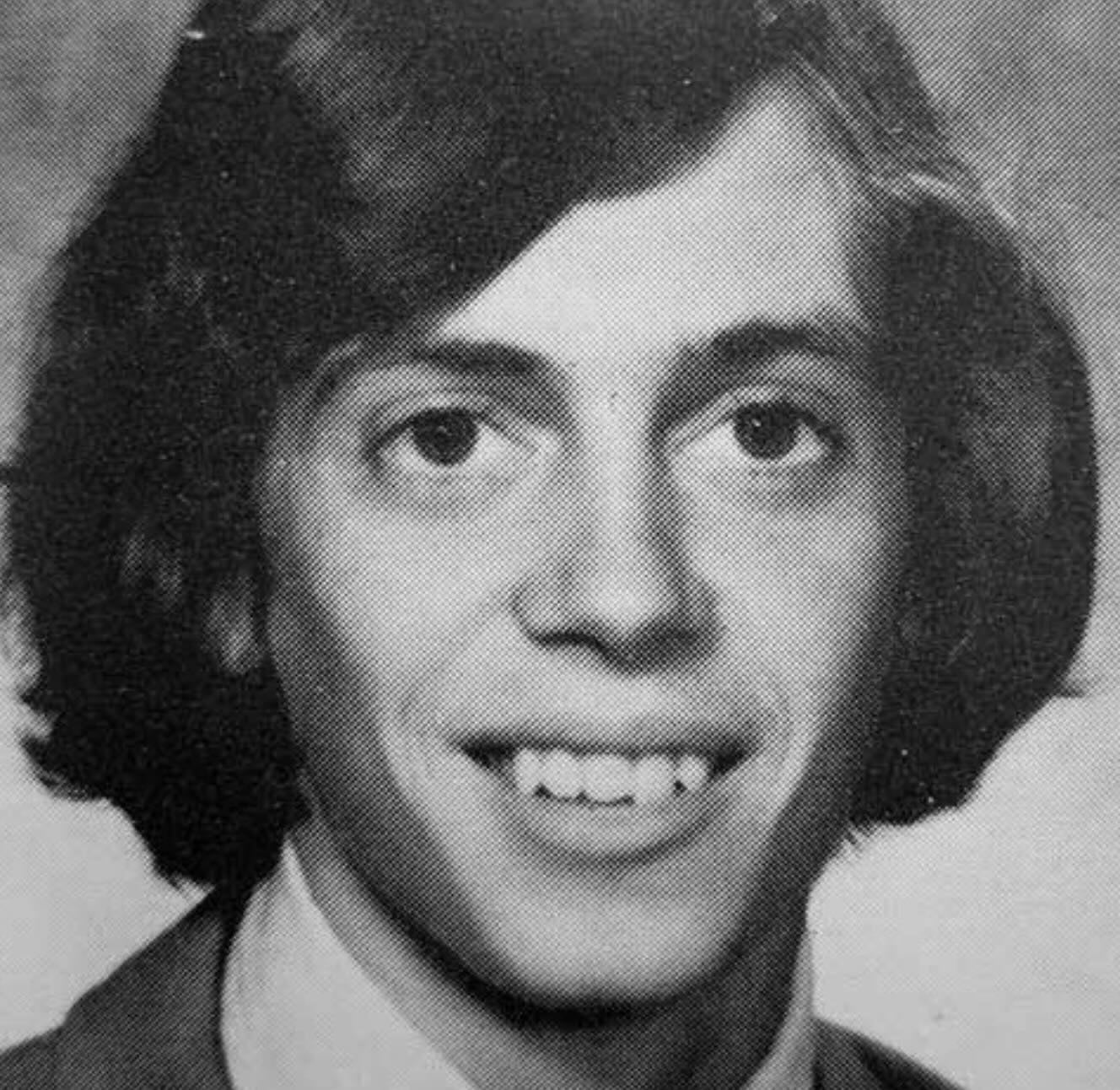 Black and white yearbook-style portrait of a young person with medium-length dark hair, wearing a suit jacket, dress shirt, and tie, smiling directly at the camera.