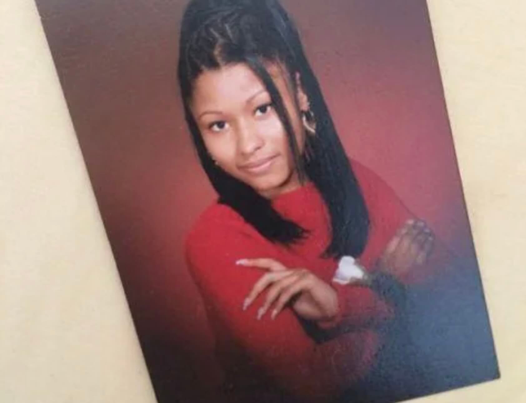 A young woman with long, straight black hair styled in a half-updo poses for a portrait. She is wearing a red sweater and has her arms crossed, smiling confidently against a plain reddish background.
