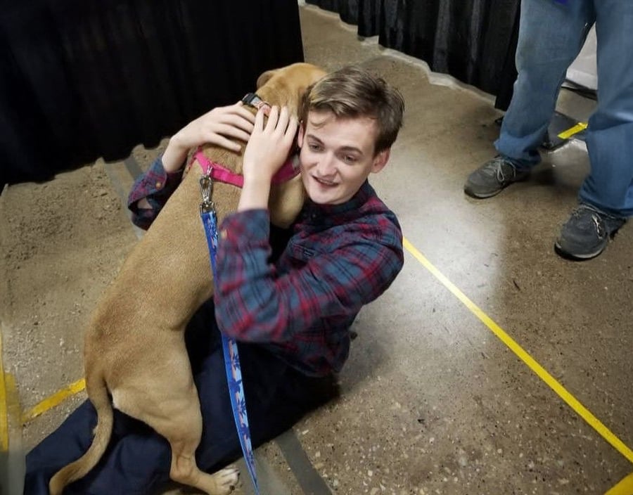 A young man sitting on the floor smiles as he hugs a tan dog with a pink collar and leash. The dog’s front paws rest on his lap. People and black curtains are visible in the background.