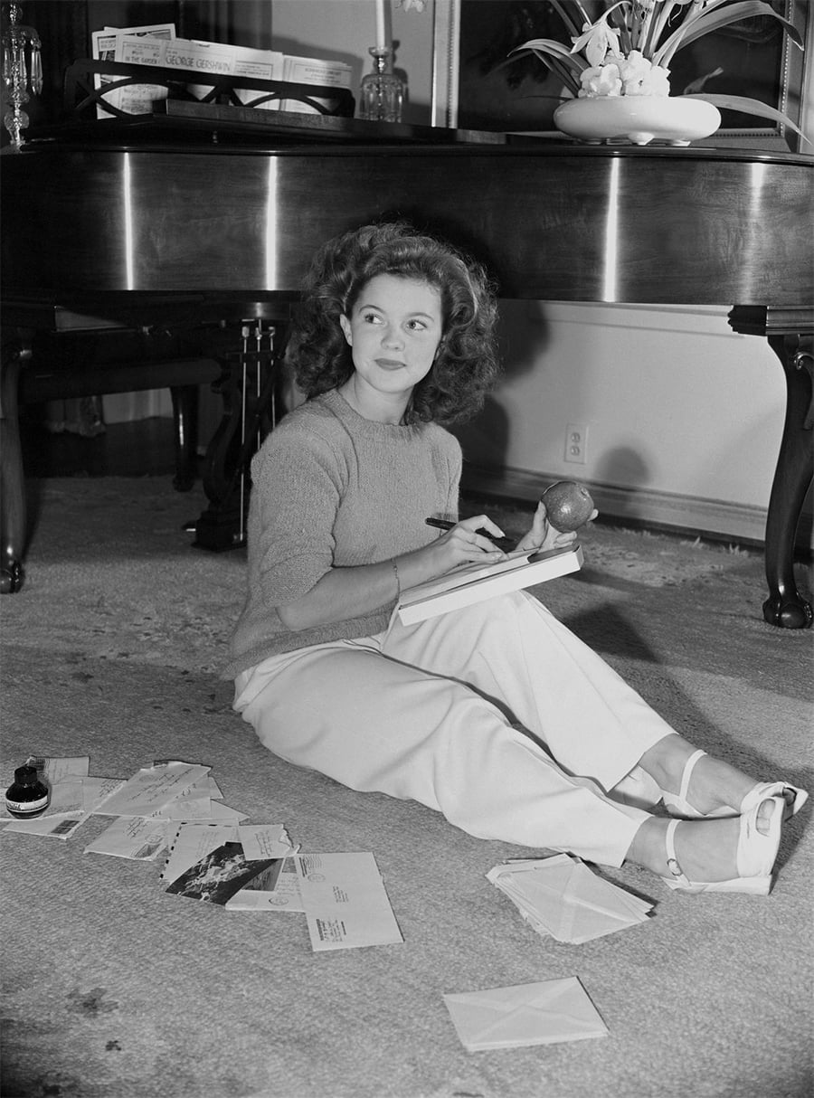 A woman sits on the floor in front of a piano, holding a notepad and pen in one hand and an apple in the other. Several opened envelopes and letters are scattered around her.
