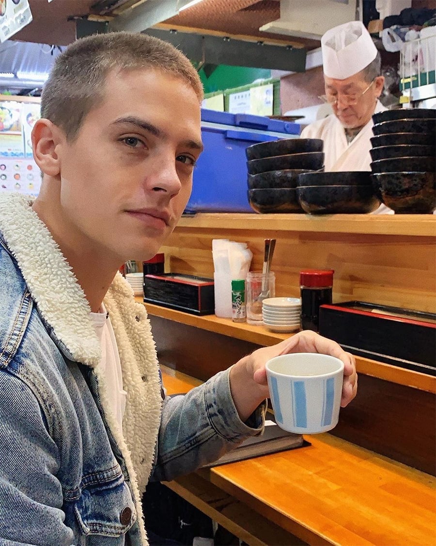 A young man in a denim jacket holds a tea cup and sits at a sushi bar counter, while a chef in a white uniform prepares food behind the counter with stacked black bowls.
