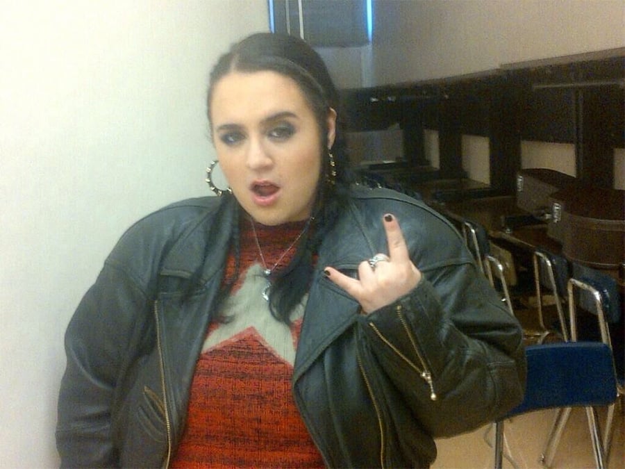 A person with dark hair, wearing a black leather jacket and hoop earrings, stands indoors making a rock-and-roll hand gesture with their mouth slightly open. There are empty chairs and guitar cases in the background.