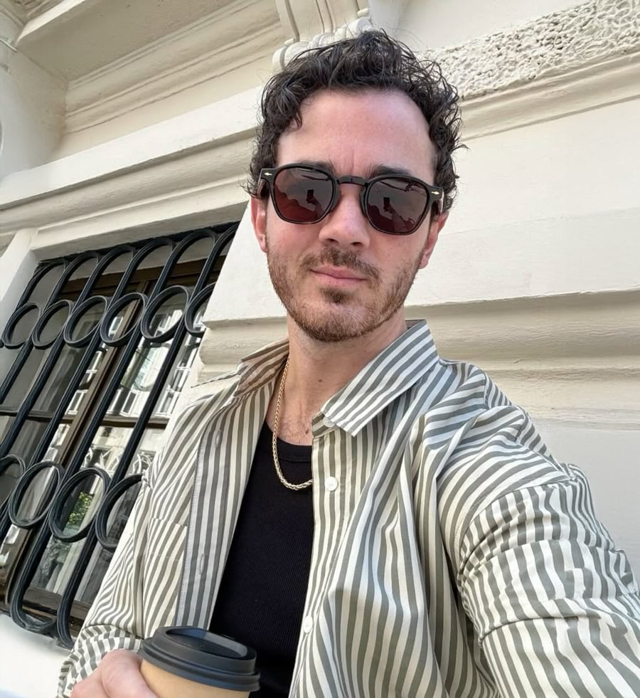 A man with curly hair wearing sunglasses, a striped button-up shirt over a black shirt, and a gold chain holds a takeaway coffee cup while standing in front of a cream-colored building with decorative details and a barred window.