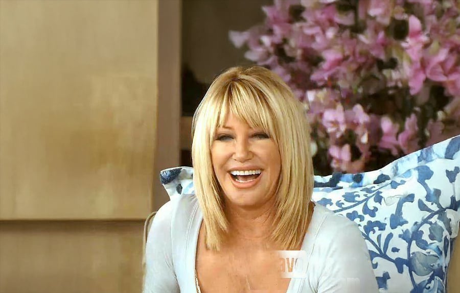 A woman with straight blonde hair smiles brightly while sitting in front of a floral arrangement and a blue-patterned pillow. She is wearing a light-colored top.