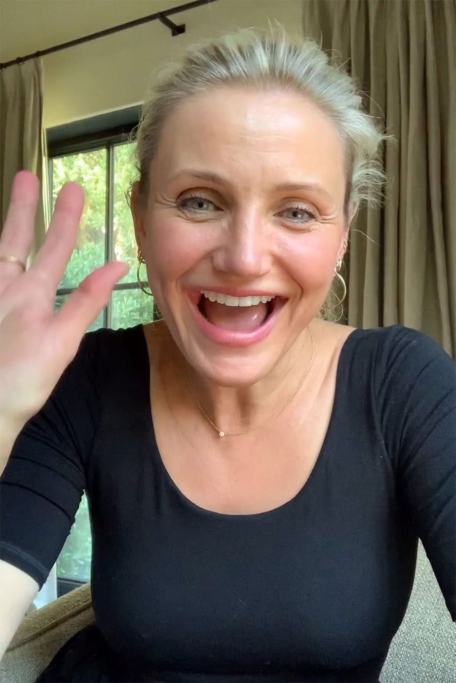 A woman with blonde hair in a black top smiles and waves at the camera. She is indoors, with beige curtains and a large window in the background.