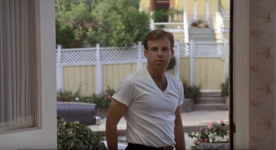 A man wearing glasses and a white T-shirt stands at an open door, looking inside. A garden, a white fence, and a staircase leading to a yellow house are visible in the background.