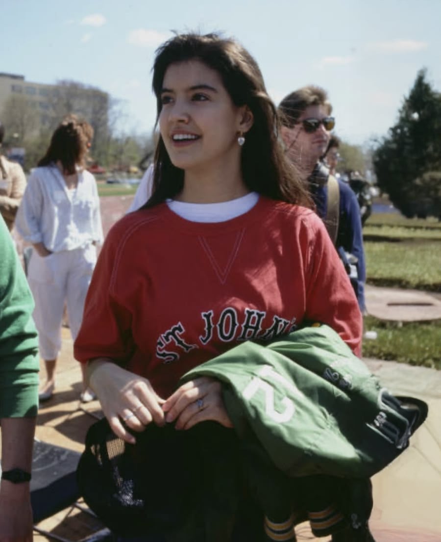 A young woman outdoors smiles while holding a green jacket. She wears a red "ST JOHN'S" sweatshirt and earrings. Other people are in the background on a sunny day.