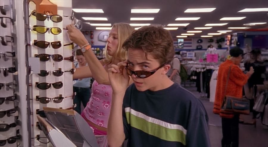 A teenage boy lowers sunglasses to look over them while standing by a sunglasses display in a store; a girl in a pink top browses in the background. The store is bright and busy with shoppers.