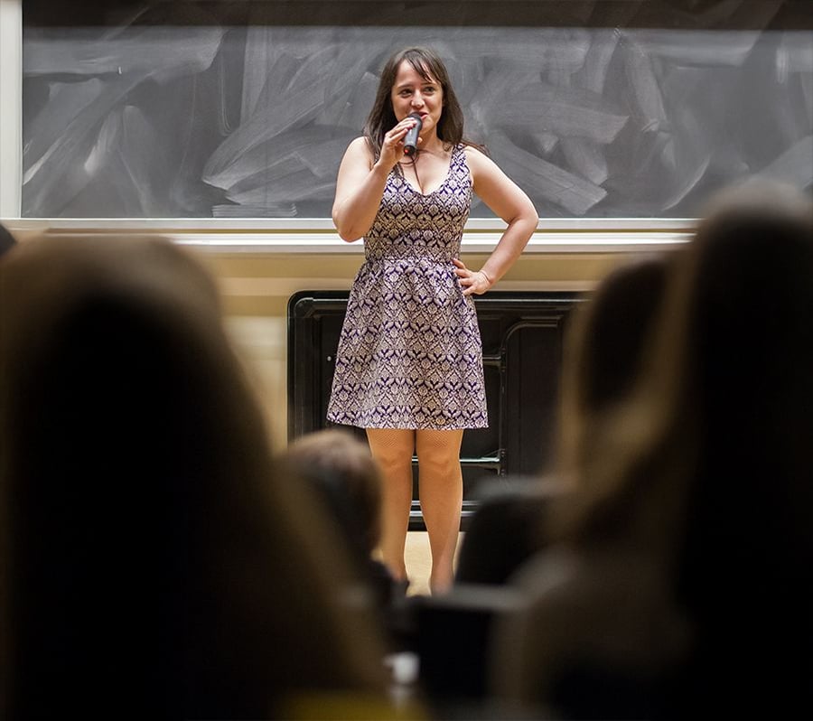 A woman in a patterned dress stands on stage with a microphone, speaking to an audience in front of a large chalkboard. She has one hand on her hip and appears confident. The audience is visible in the foreground.