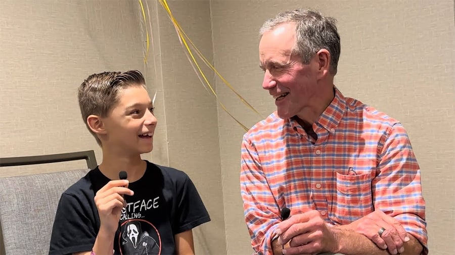 A smiling older man in a plaid shirt talks with a young boy in a black graphic tee. Both are holding microphones and looking at each other, appearing to enjoy their conversation. Yellow balloons are in the background.