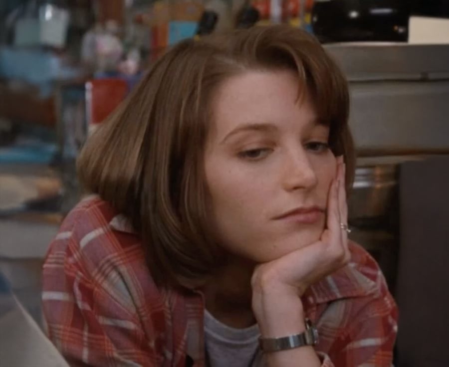 A woman with short brown hair wearing a red plaid shirt rests her head on her hand, looking thoughtful or bored. The background appears to be a kitchen or diner with various items visible.