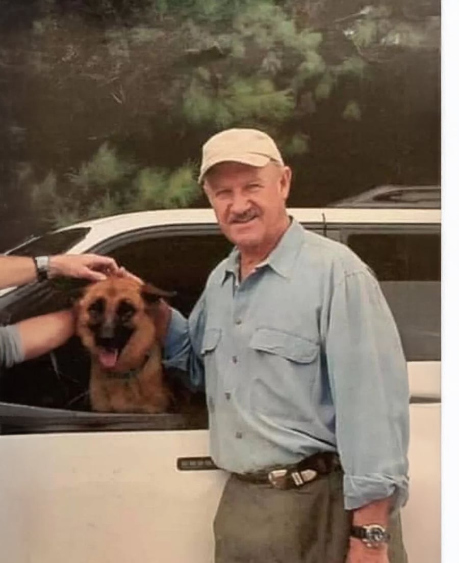 An older man in a light blue shirt and beige cap stands next to a white vehicle, smiling, while a brown dog with its tongue out looks out from the open car window. Trees are visible in the background.