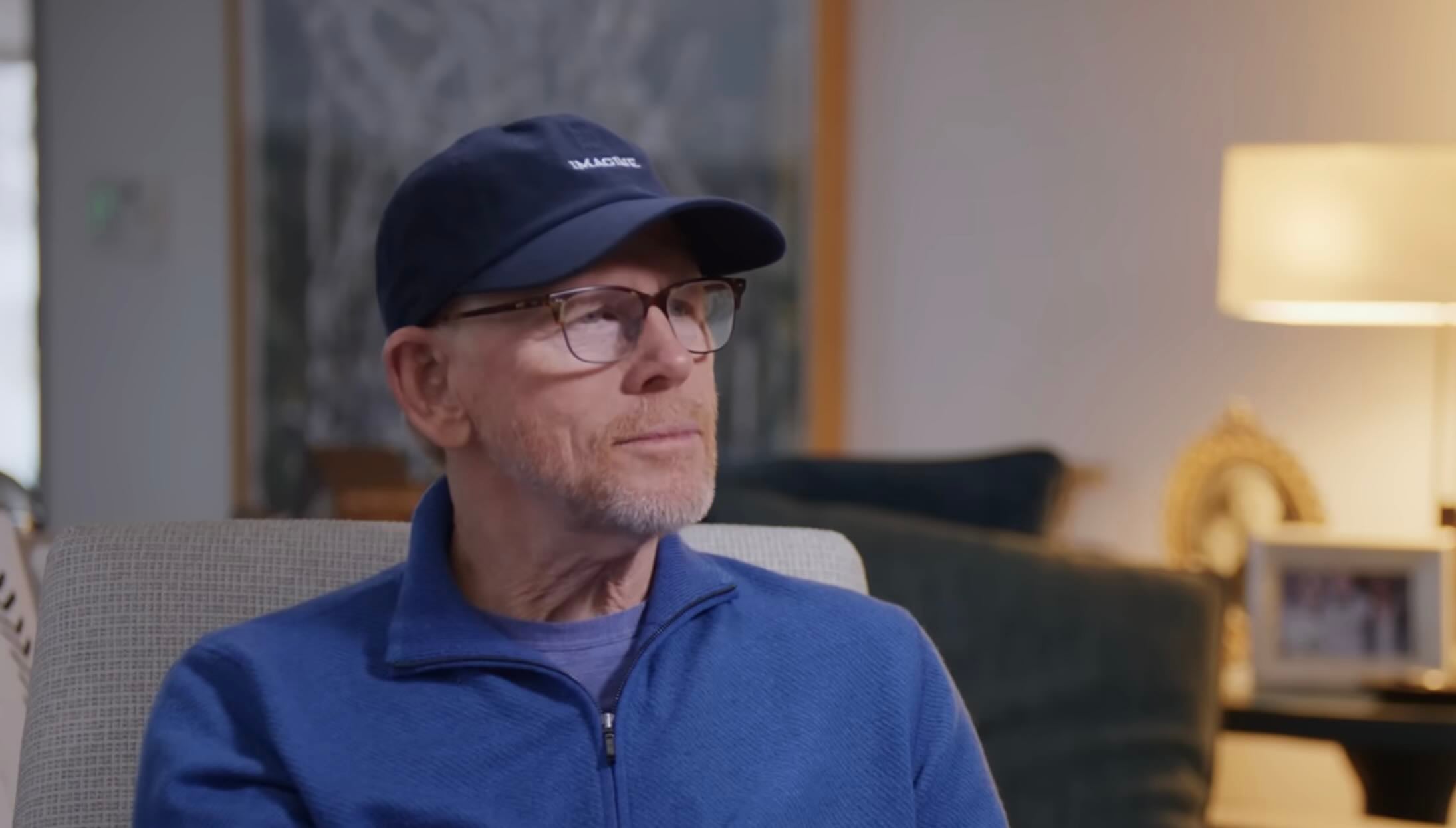 A man wearing a navy blue cap, glasses, and a blue zip-up sweater sits on a beige chair in a softly lit living room, with a blurred background featuring a lamp, framed photo, and a couch.