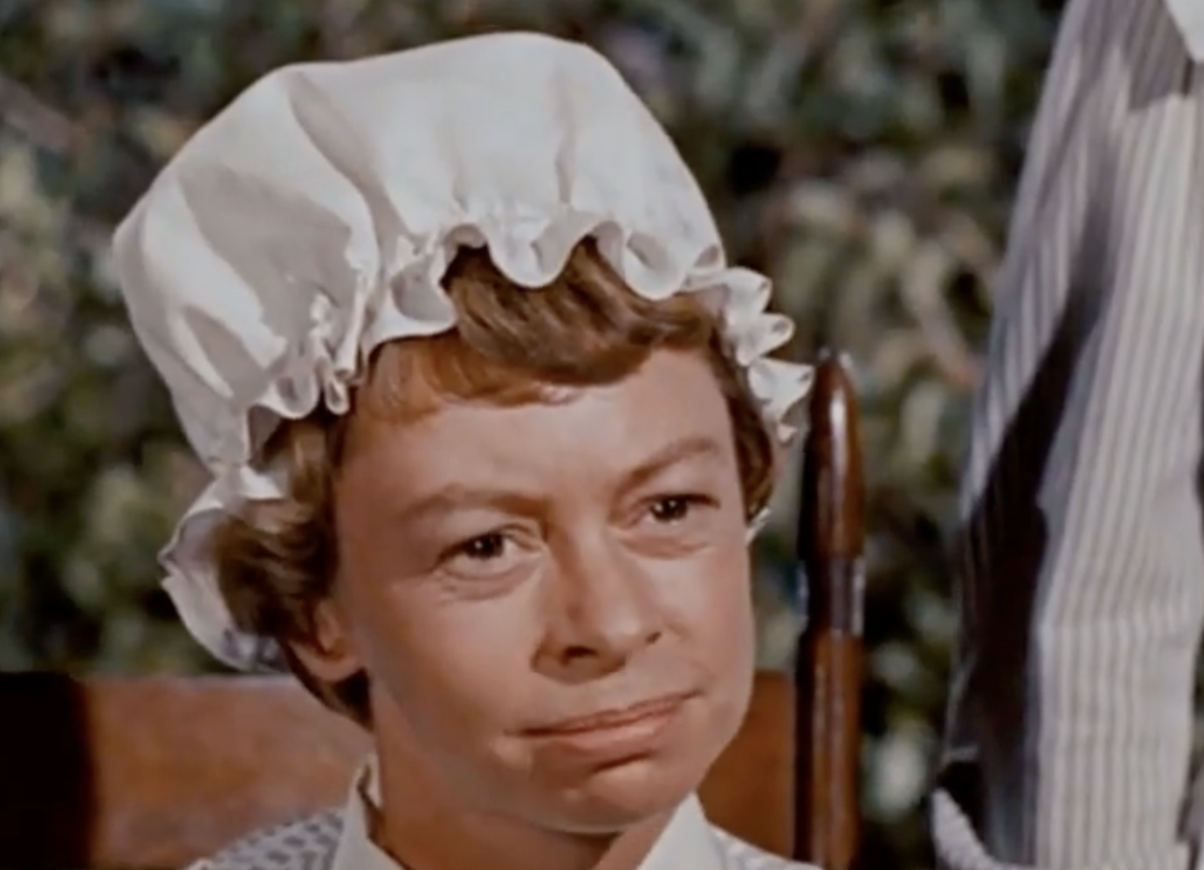 A woman wearing a white bonnet and period clothing looks slightly to the side with a neutral expression, seated outdoors with greenery in the background.