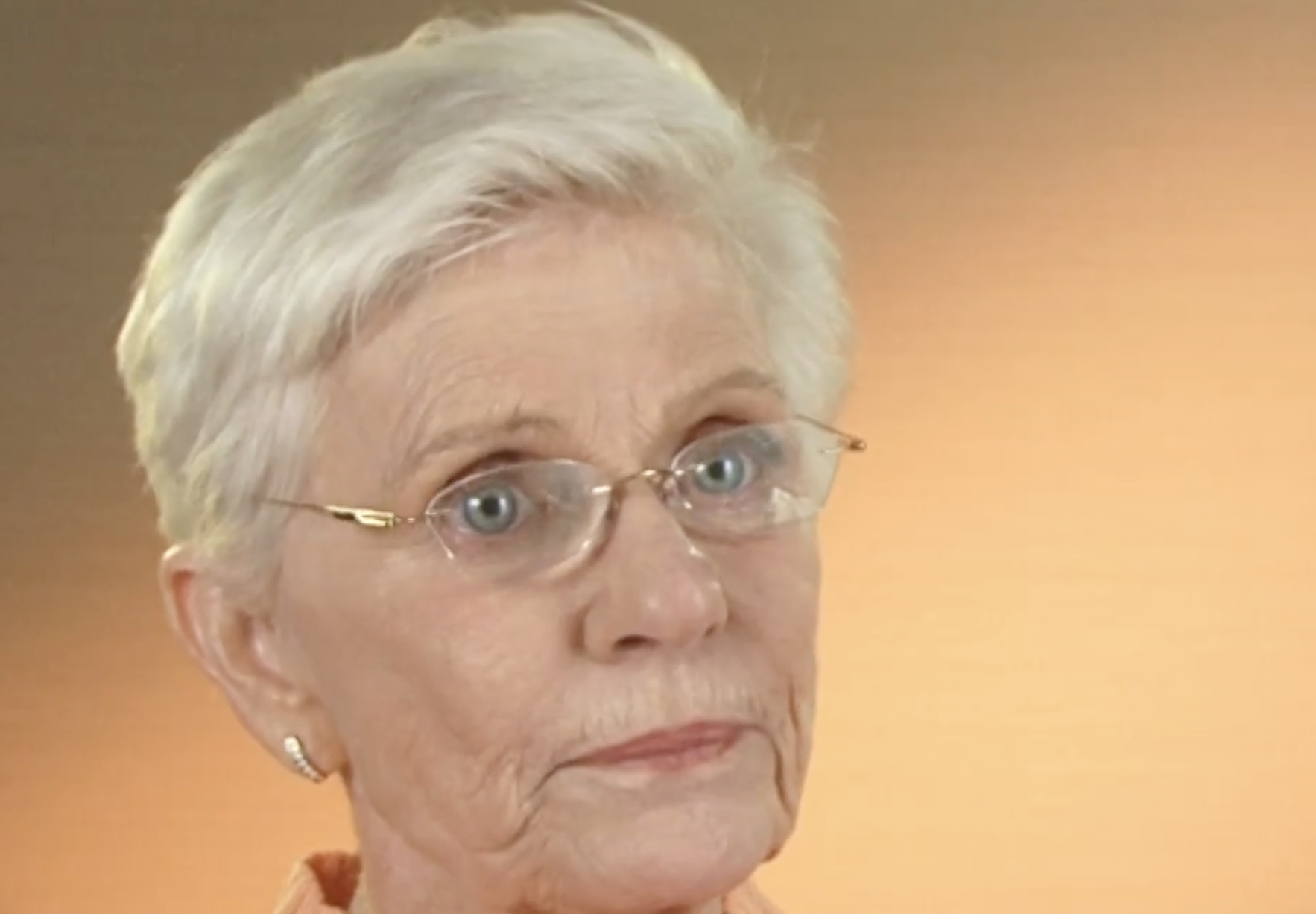 An elderly woman with short, white hair and blue eyes is wearing glasses and stud earrings. She is gazing slightly to the side with a neutral expression. The background is a soft, warm gradient.