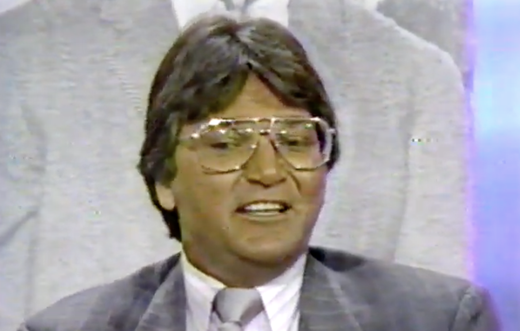 A man with brown hair and large glasses, dressed in a gray suit and tie, smiles while sitting indoors. The background appears to show a large, faded black-and-white image of a person in similar attire.